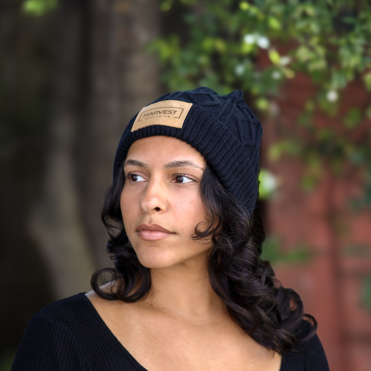 A woman wearing a dark navy blue cable-knit beanie with a tan leather patch that reads 'CABINET' on the front, photographed outdoors with a blurred background of green foliage and a brick wall.