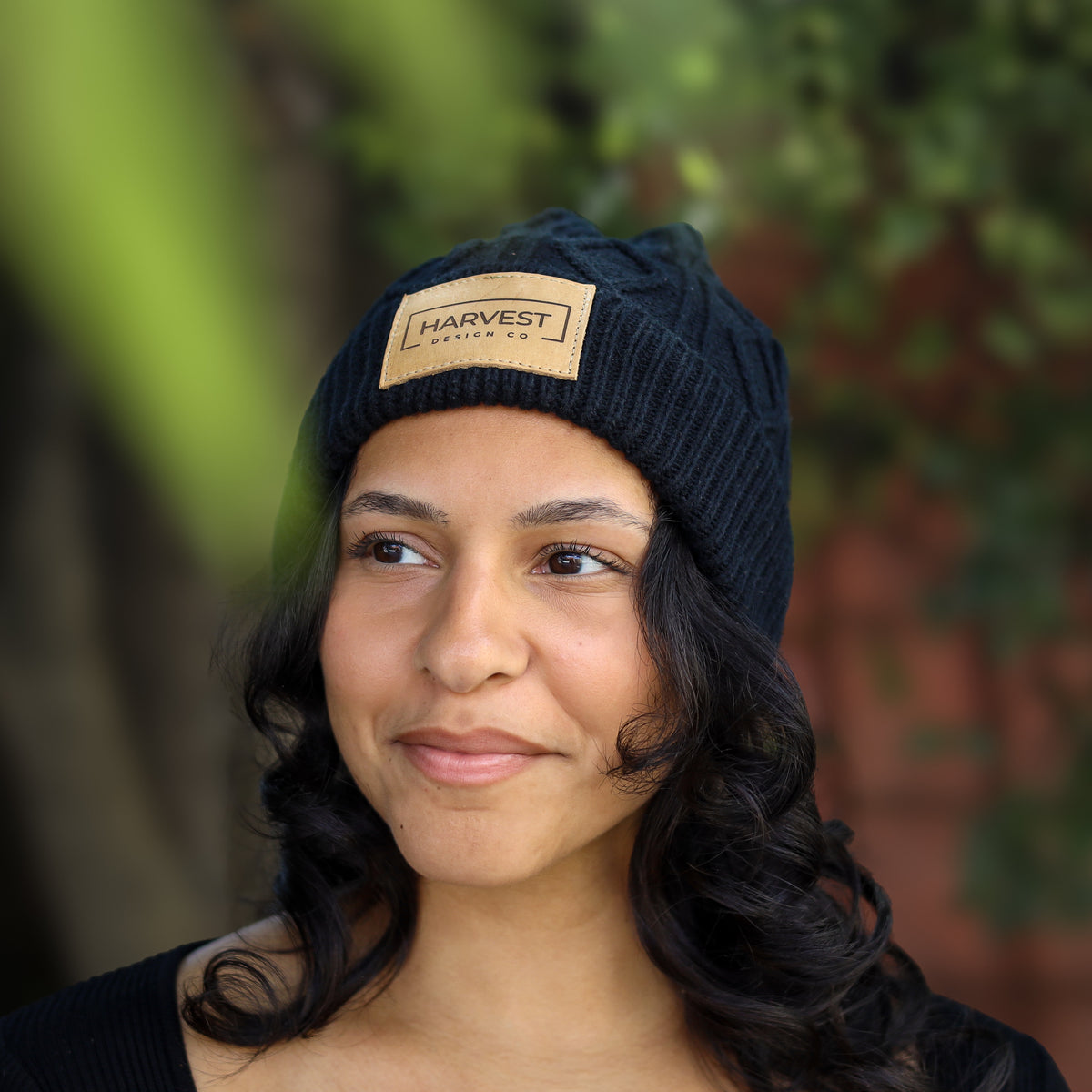 A woman wearing a dark navy blue cable-knit beanie with a tan rectangular patch that reads 'HARVEST DESIGN CO.' in black text, photographed outdoors with blurred greenery and a brick wall in the background.