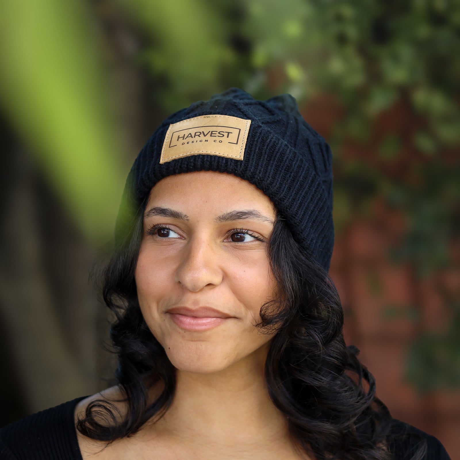A woman wearing a dark navy blue cable-knit beanie with a tan rectangular patch that reads 'HARVEST DESIGN CO.' in black text, photographed outdoors with blurred greenery and a brick wall in the background.