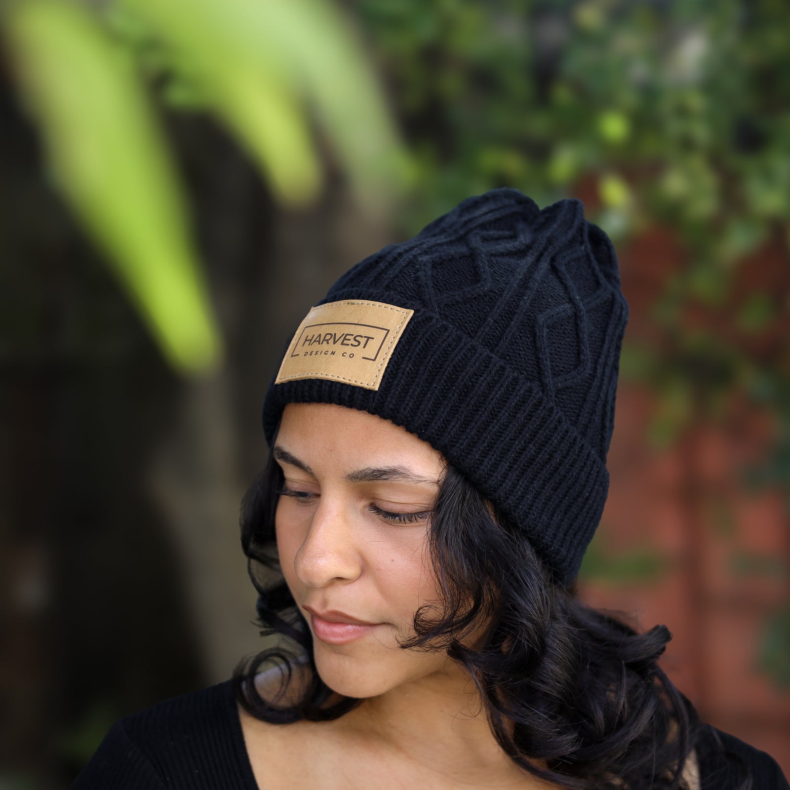 A woman wearing a black cable-knit beanie with a tan leather patch that reads 'HARVEST CLOTHING CO.' in black text, photographed outdoors with a blurred green and brown background.