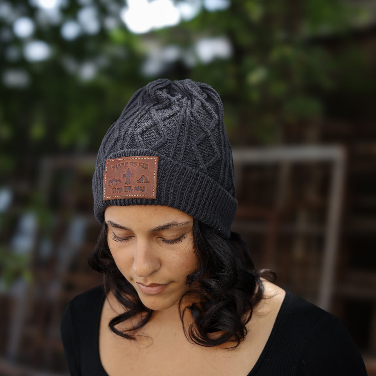 A woman wearing a dark gray cable-knit beanie with a brown leather patch on the front that reads 'PETER PO ACE' and 'EST. 2014'.