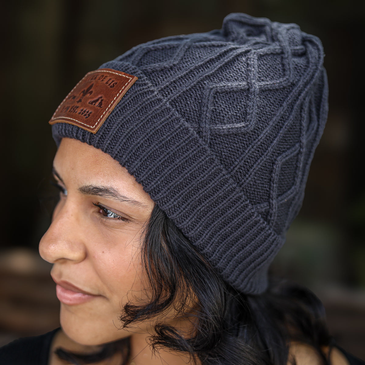 A close-up side profile of a woman wearing a dark gray cable-knit beanie with a brown leather patch on the side.