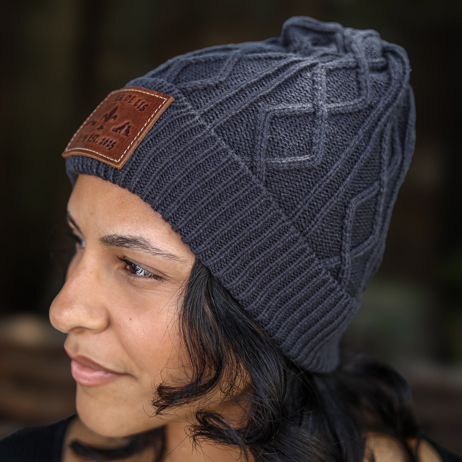 A close-up side profile of a woman wearing a dark gray cable-knit beanie with a brown leather patch on the side.