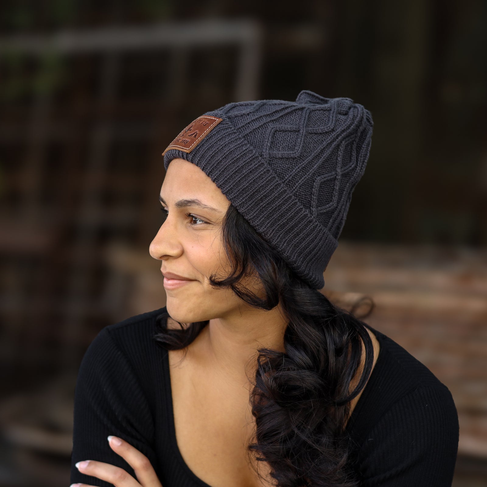 A woman wearing a dark gray cable-knit beanie with a brown leather patch on the side, photographed outdoors against a blurred background.